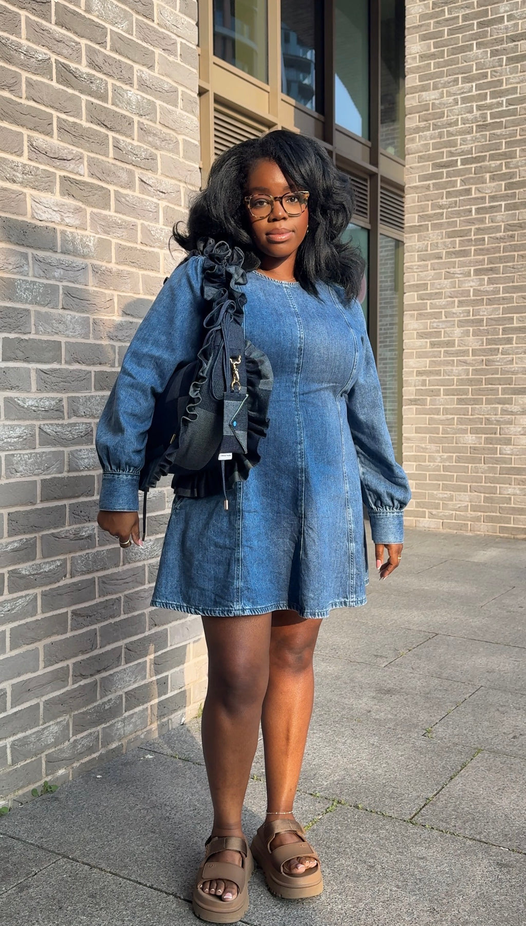 Woman wearing a blue denim dress holding a denim ruffled crescent bag  standing against a brick wall.