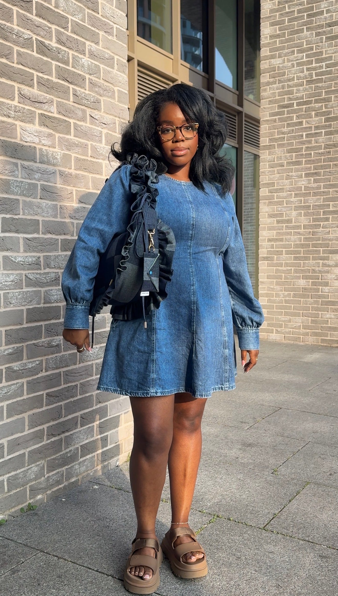 Woman wearing a blue denim dress holding a denim ruffled crescent bag  standing against a brick wall.