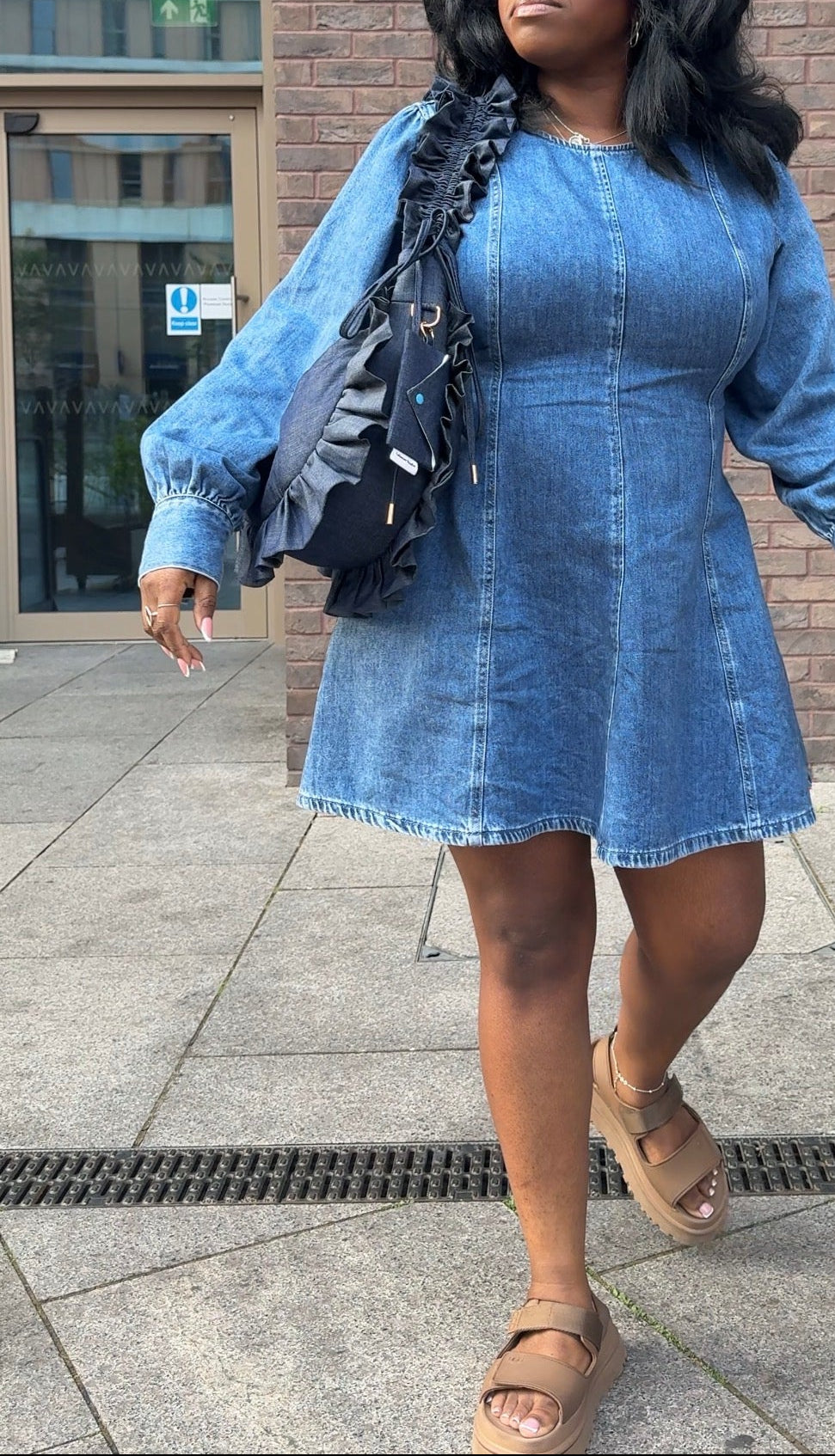 Woman wearing a denim dress holding a ruffled, denim crescent bag standing on a sidewalk with a building in the background