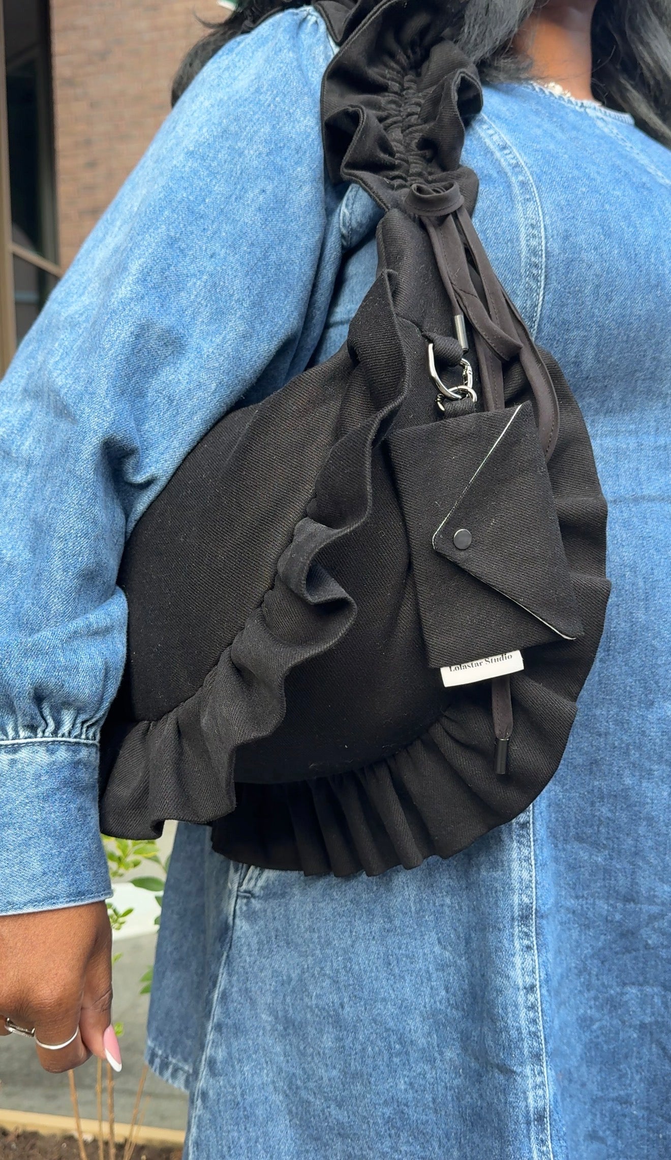 Black denim ruffled crescent  bag worn over one shoulder by a person wearing a blue denim dress.