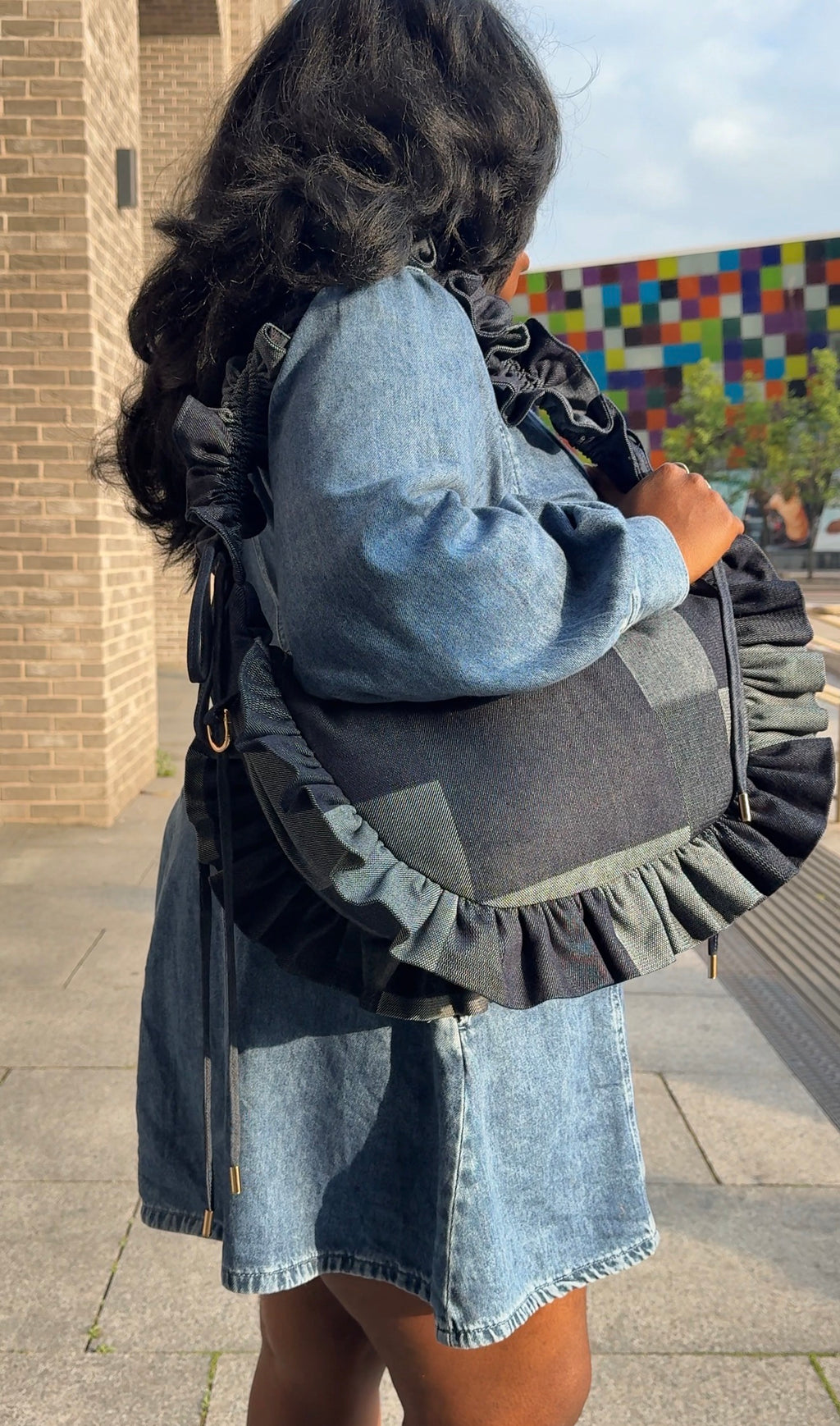 Person wearing a denim dress and  a denim ruffled crescent bag  on a sidewalk.