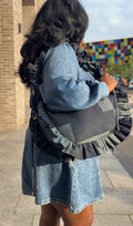 Person wearing a denim dress and  a denim ruffled crescent bag  on a sidewalk.