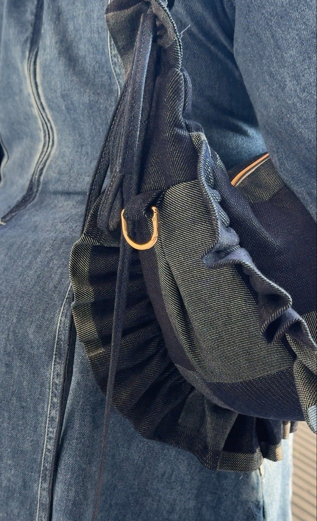 Close-up of a denim ruffled crescent bag  with a gold buckle on a person wearing a denim dress.
