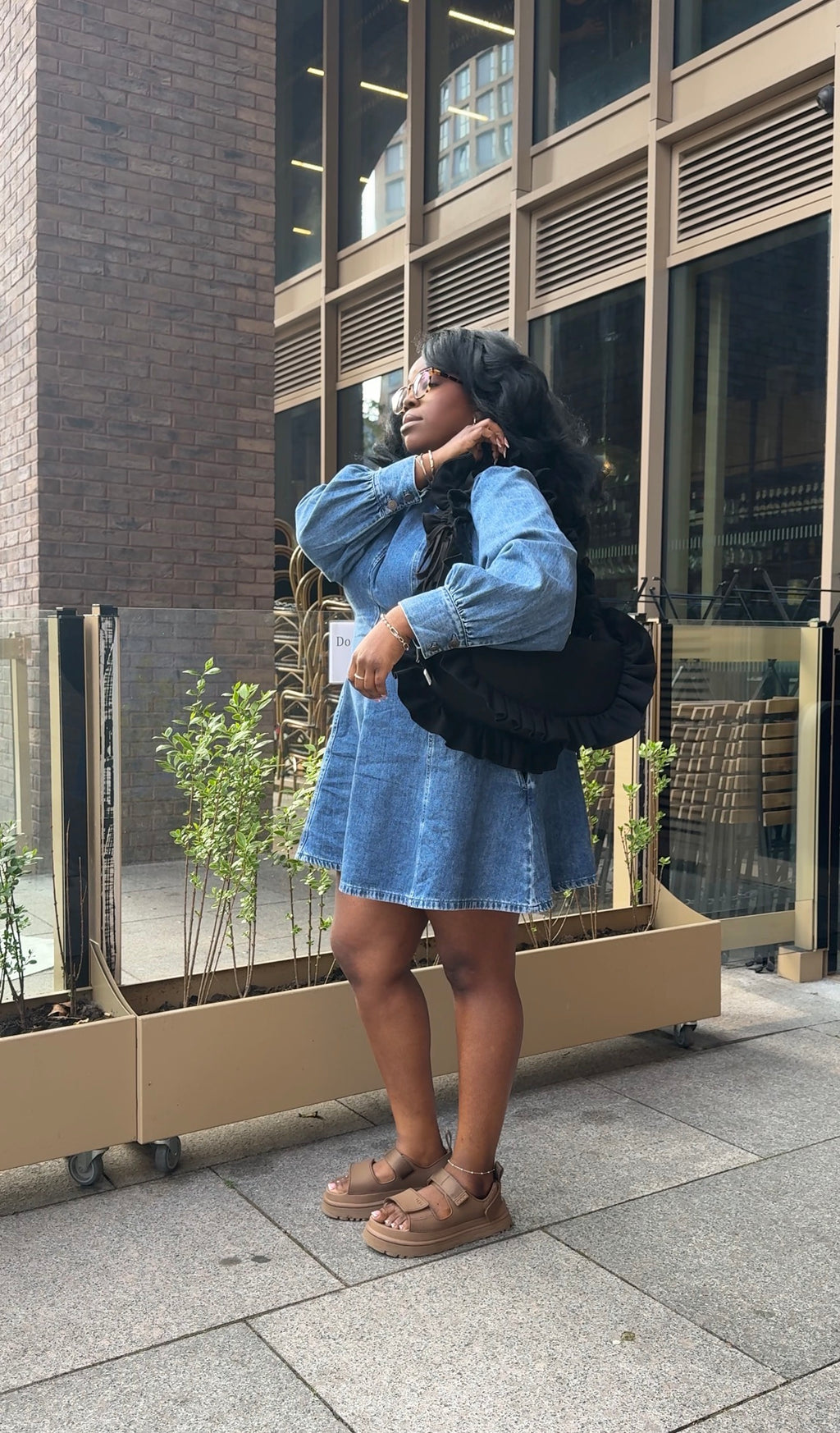 Person wearing a denim dress and a denim ruffled crescent bag  standing on a sidewalk with a building in the background