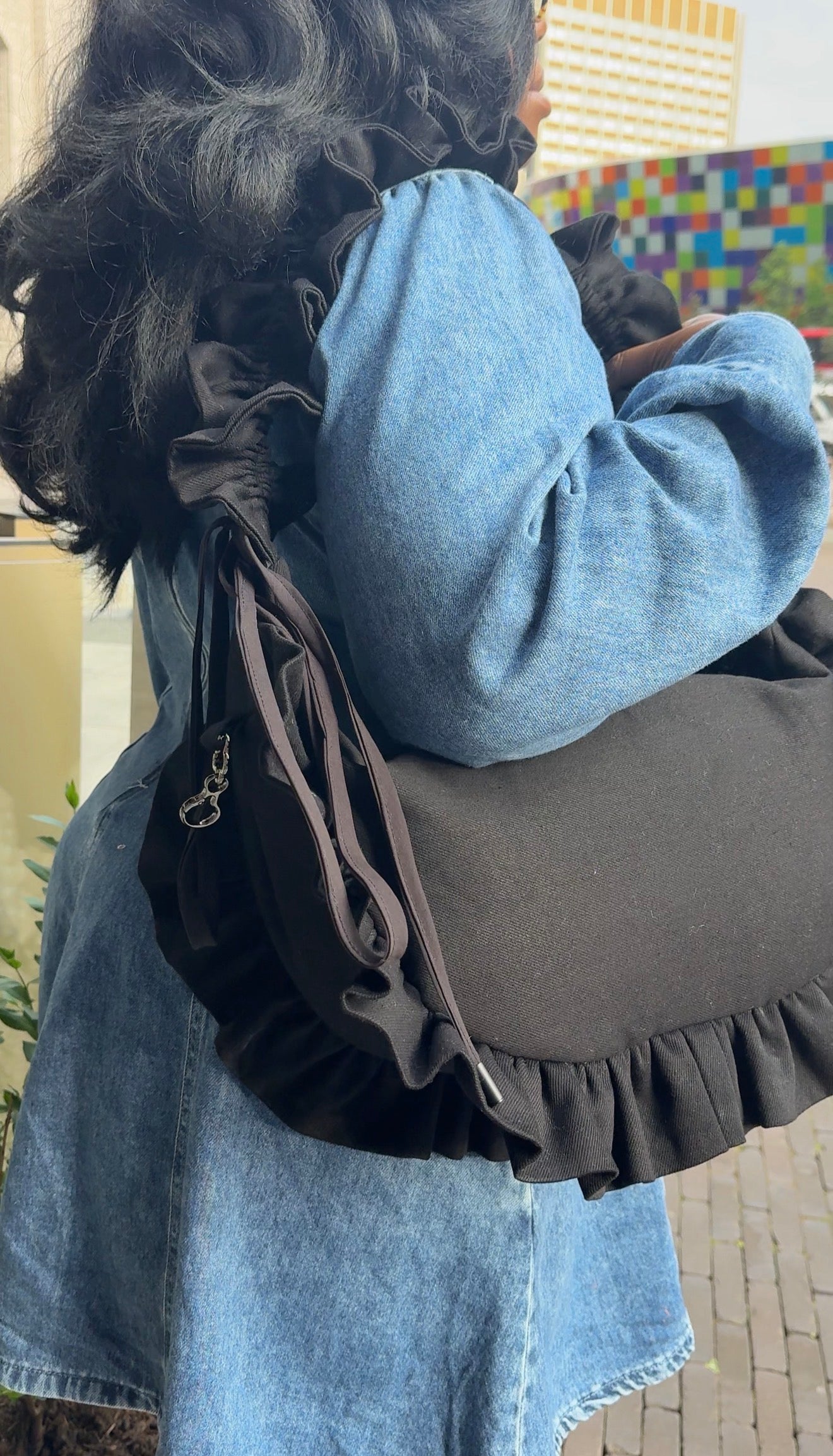 Person wearing a black denim ruffled crescent bag  bag with a colourful mural in the background