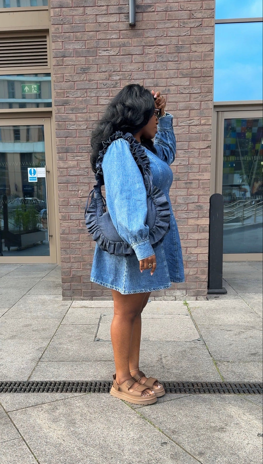 Woman in a denim dress holding a ruffled crescent handbag standing on a sidewalk with a brick building in the background