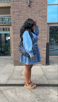 Woman in a denim dress holding a ruffled crescent handbag standing on a sidewalk with a brick building in the background