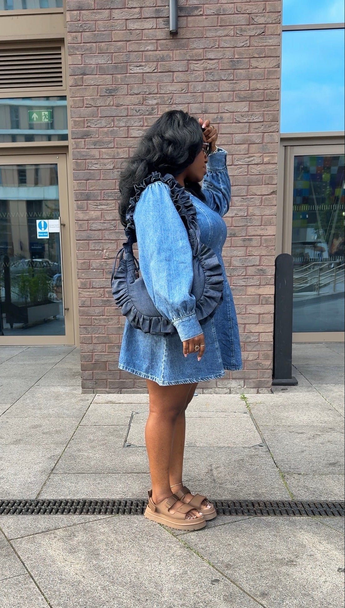 Woman in a denim dress holding a ruffled crescent handbag standing on a sidewalk with a brick building in the background