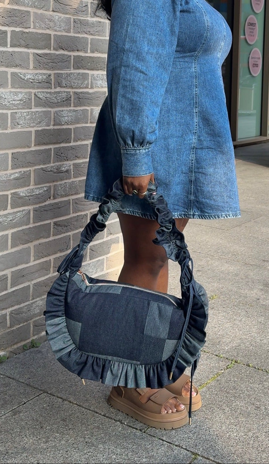 Woman in a denim dress holding a denim ruffled crescent  handbag against a brick wall.