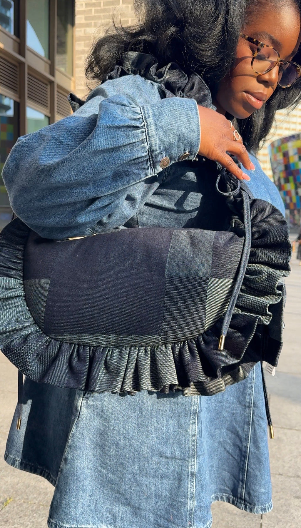 Person wearing a denim jacket and holding a black and gray denim ruffled crescent bag 