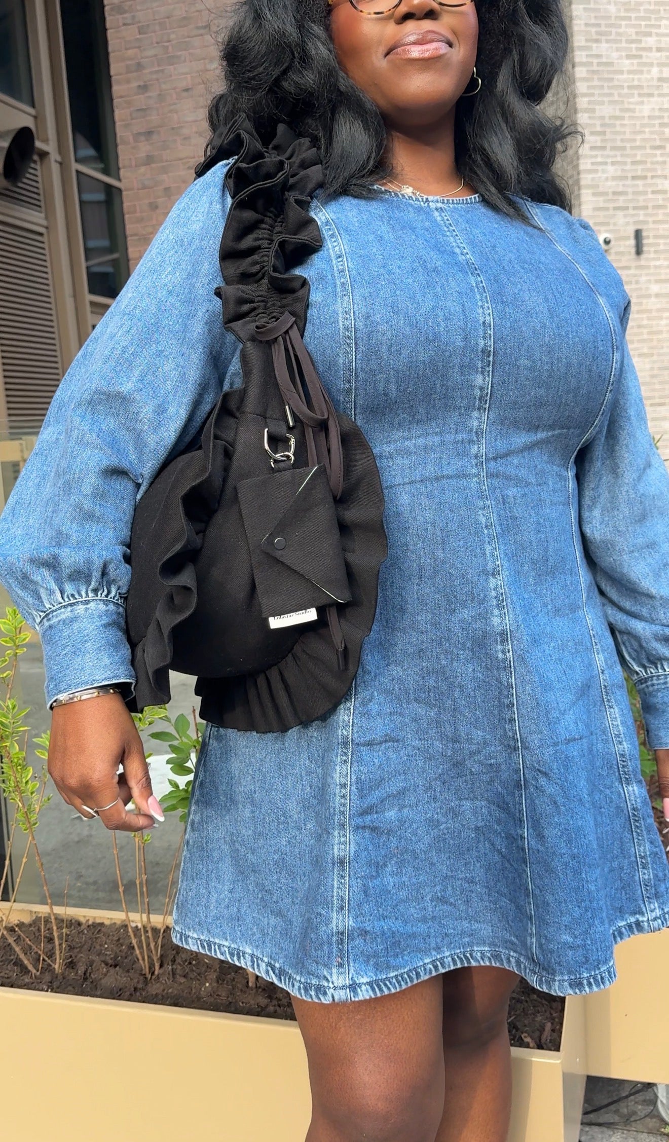 Woman wearing a denim dress with a black denim ruffled crescent bag  outdoors.