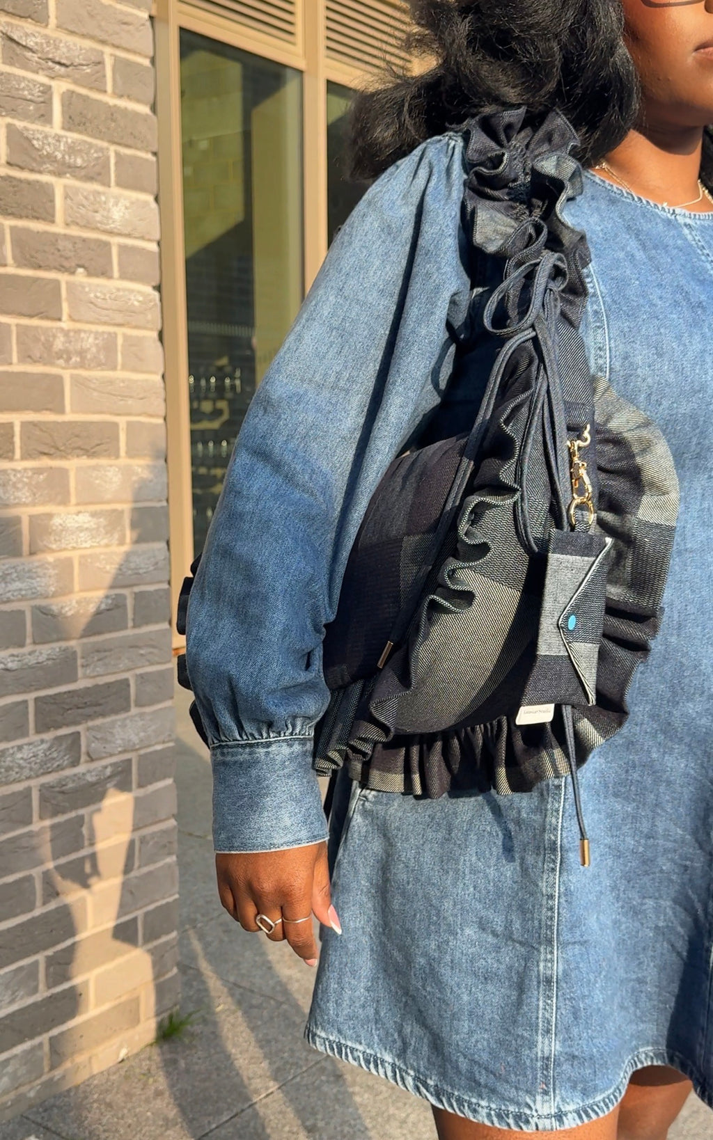 Person wearing a denim jacket and skirt with a denim ruffled crescent bag  against a brick wall.