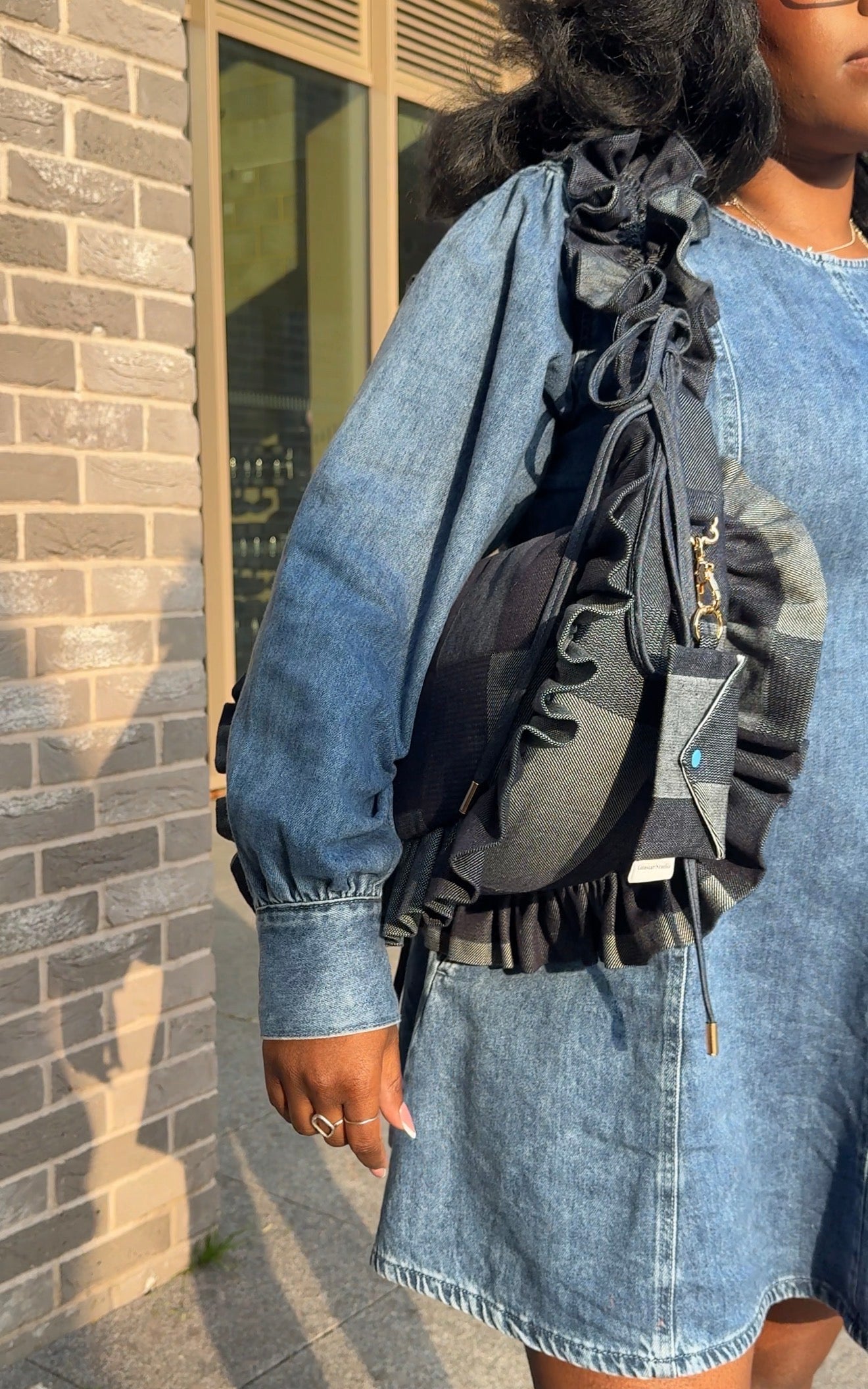 Person wearing a denim jacket and skirt with a denim ruffled crescent bag  against a brick wall.
