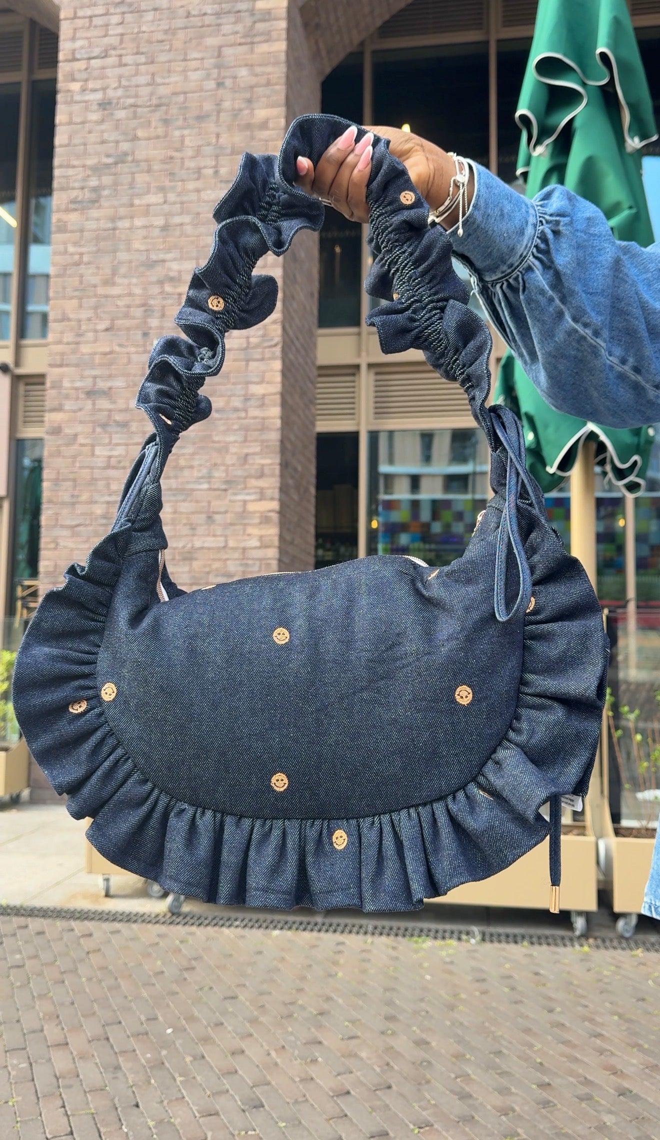 Person holding a blue denim ruffled crescent handbag  with ruffled details outdoors.