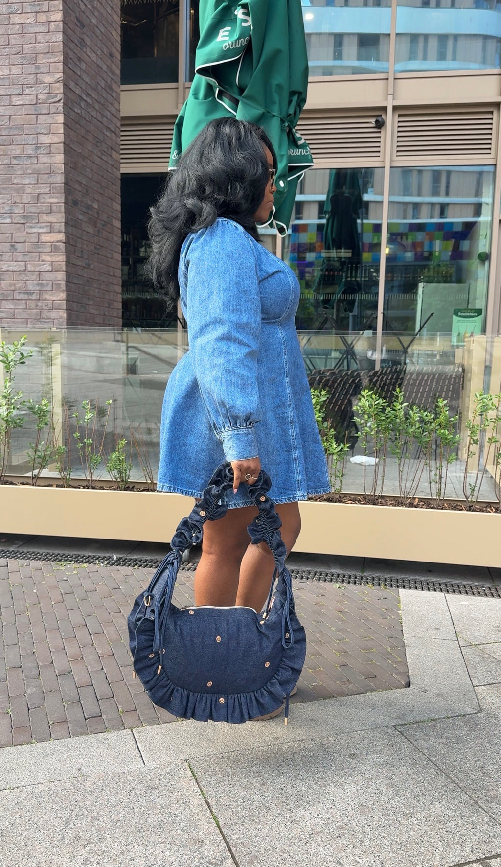 Woman in a blue dress holding a denim ruffled crescent bag standing on a street with a building in the background