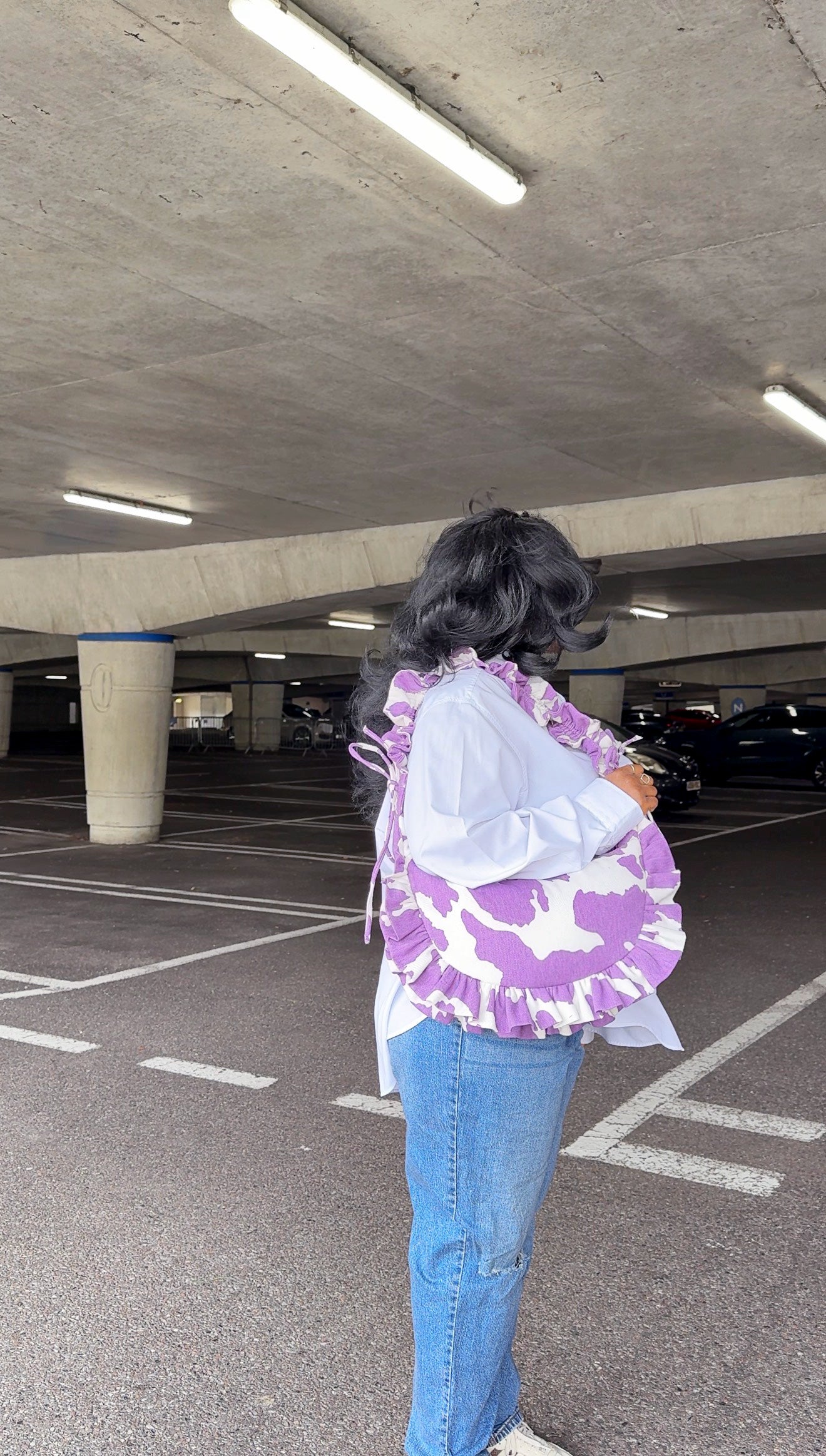 Person holding a purple amethyst cow print ruffled handbag and blue jeans standing in an underground parking lot.