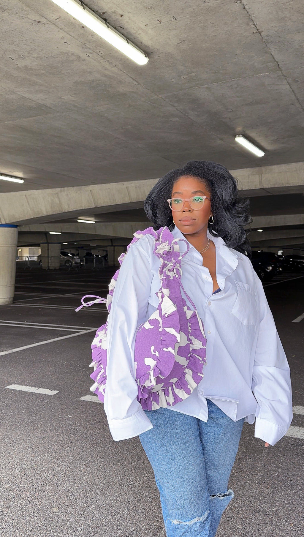 Person wearing a white shirt and blue jeans with a purple amethyst cow print ruffled handbag in an underground parking lot.