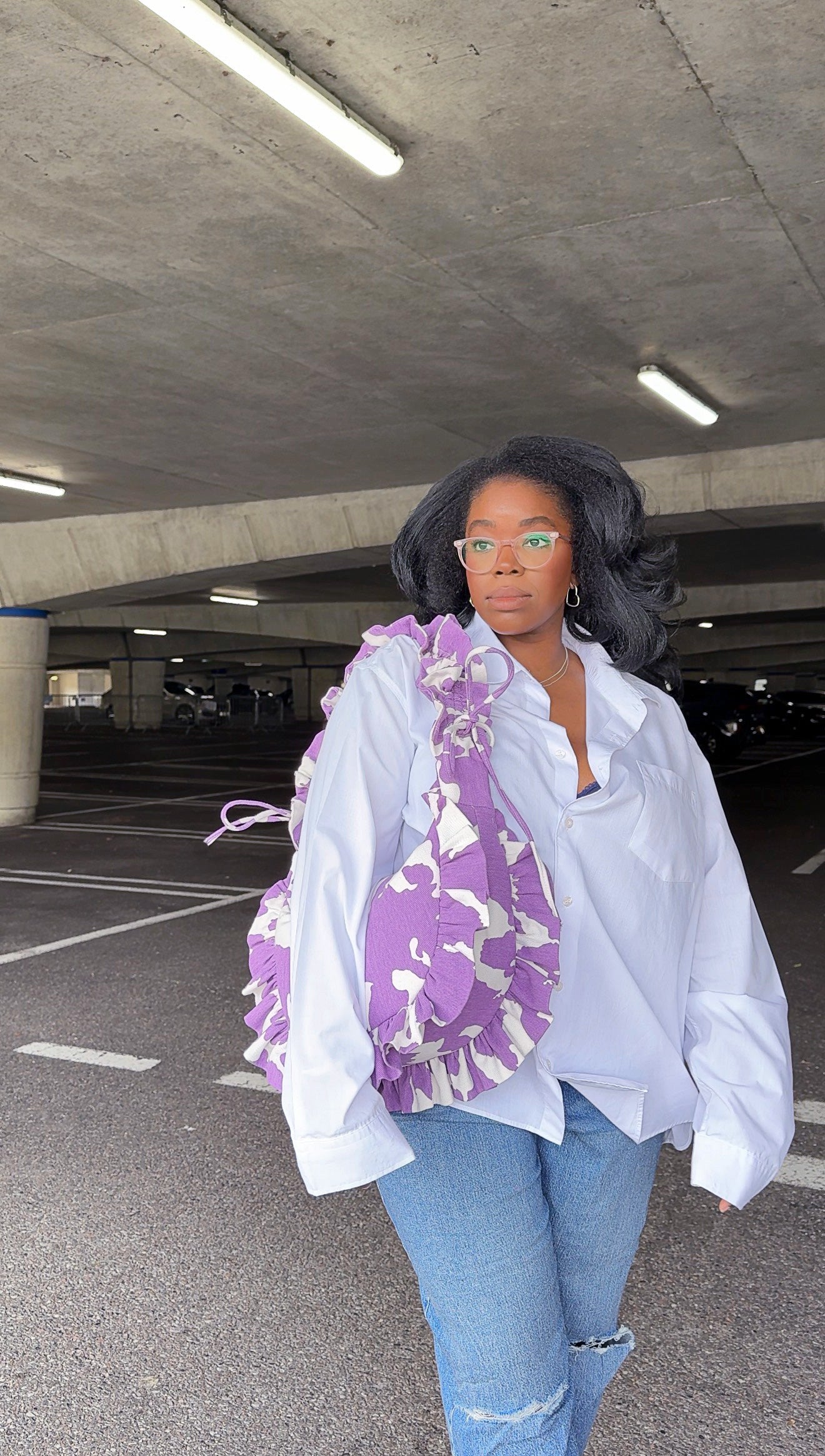 Person wearing a white shirt and blue jeans with a purple amethyst cow print ruffled handbag in an underground parking lot.