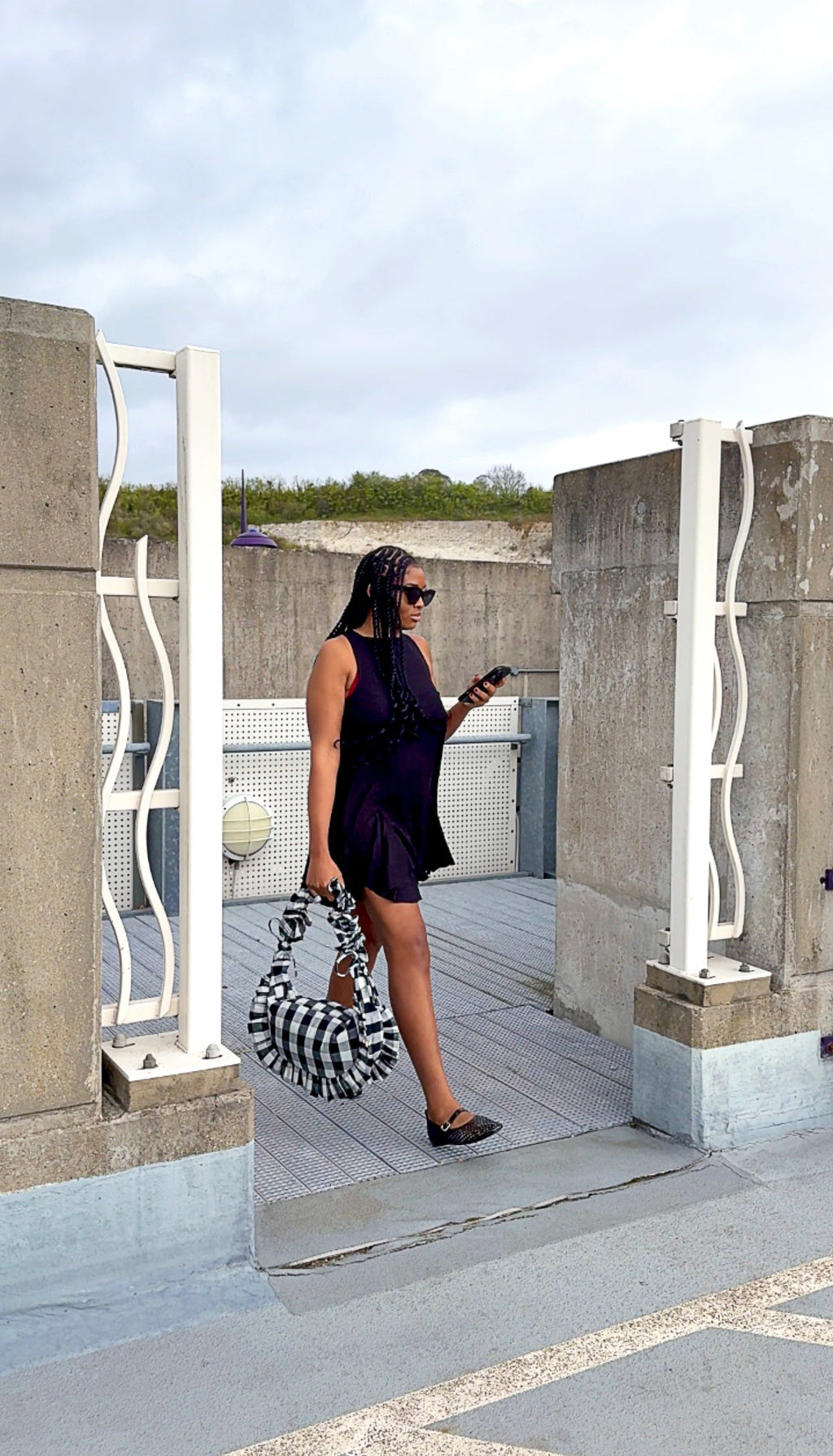 Woman in a black dress standing on a rooftop with concrete walls and a cloudy sky, holding a black and white checkered ruffle crescent bag