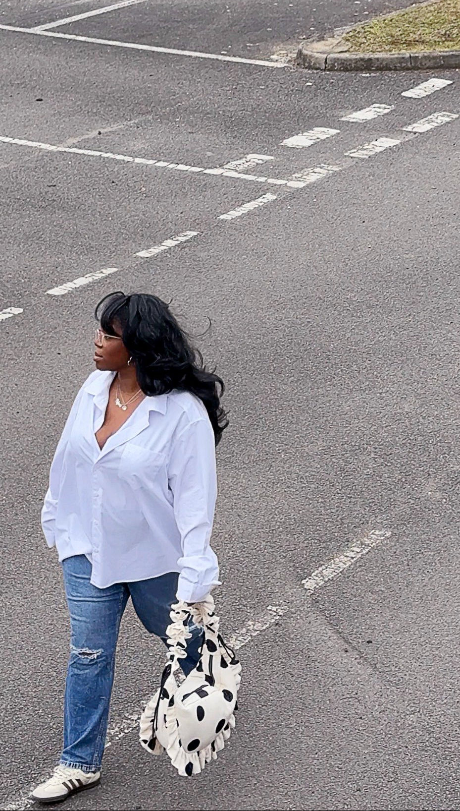 Woman in a white shirt and jeans standing in a parking lot with cones, holding a white polka dot crescent handbag.