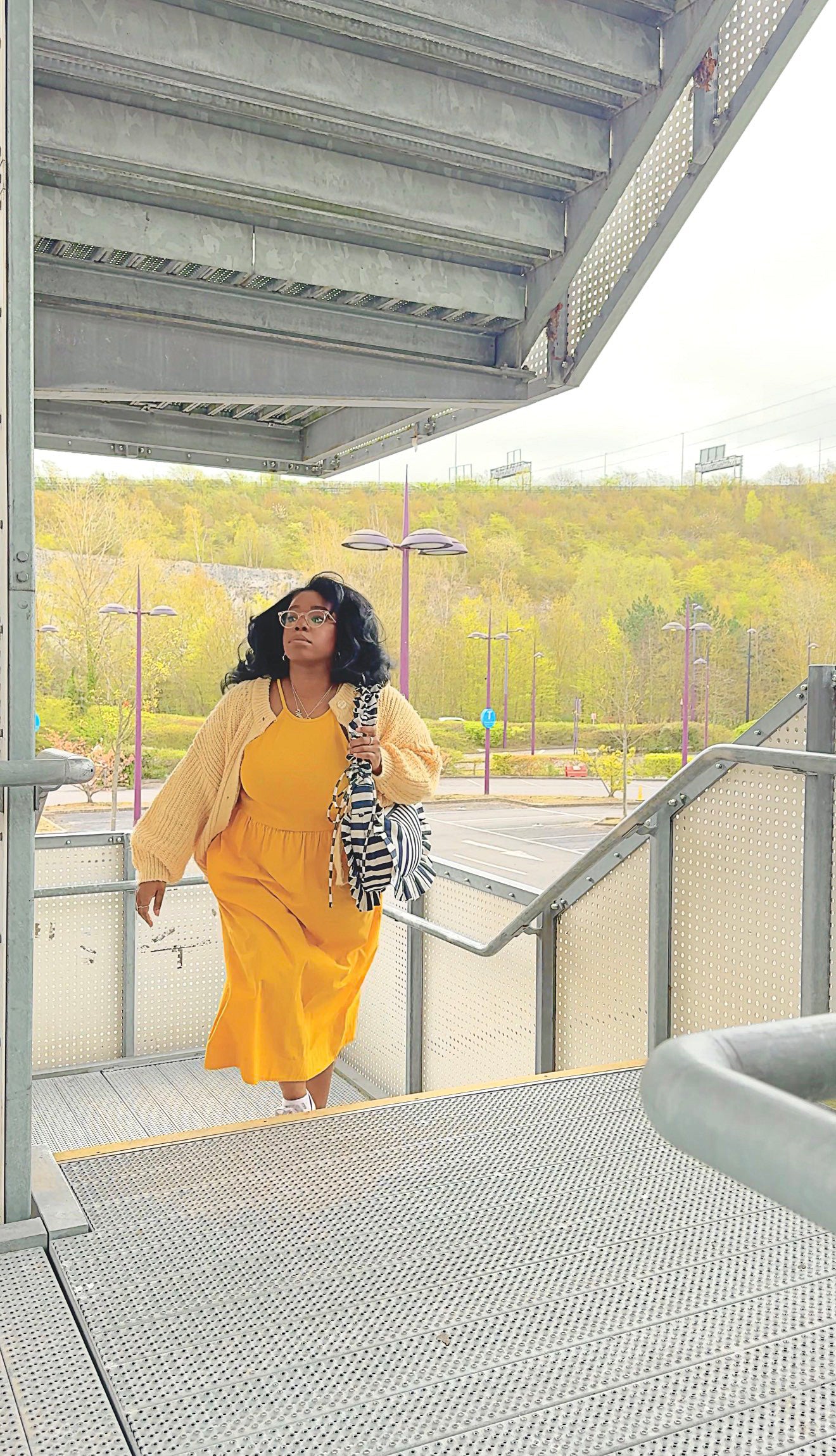 person walking up parking lot stairs holding navy and white striped ruffled crescent bag styled with a bright yellow dress. 