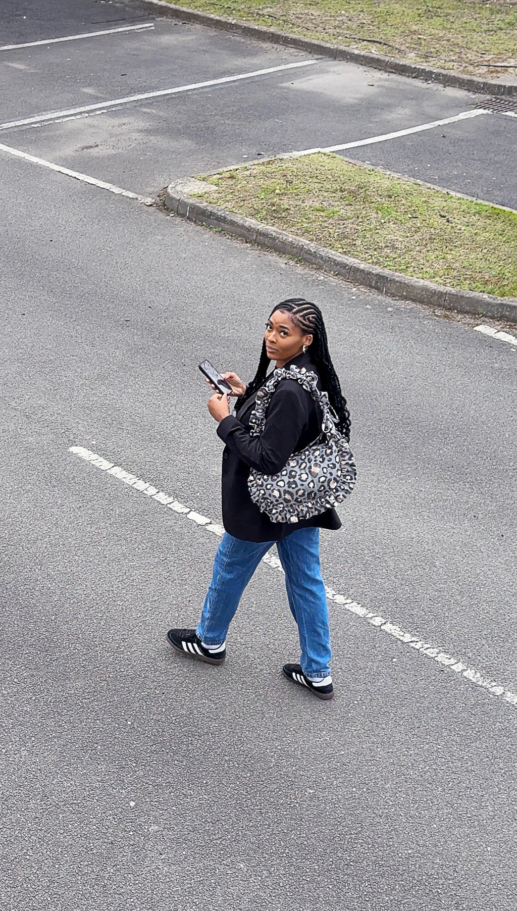 Person walking on a road holding a phone and wearing a slate leopard print ruffled crescent bag.