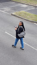 Person walking on a road holding a phone and wearing a slate leopard print ruffled crescent bag.