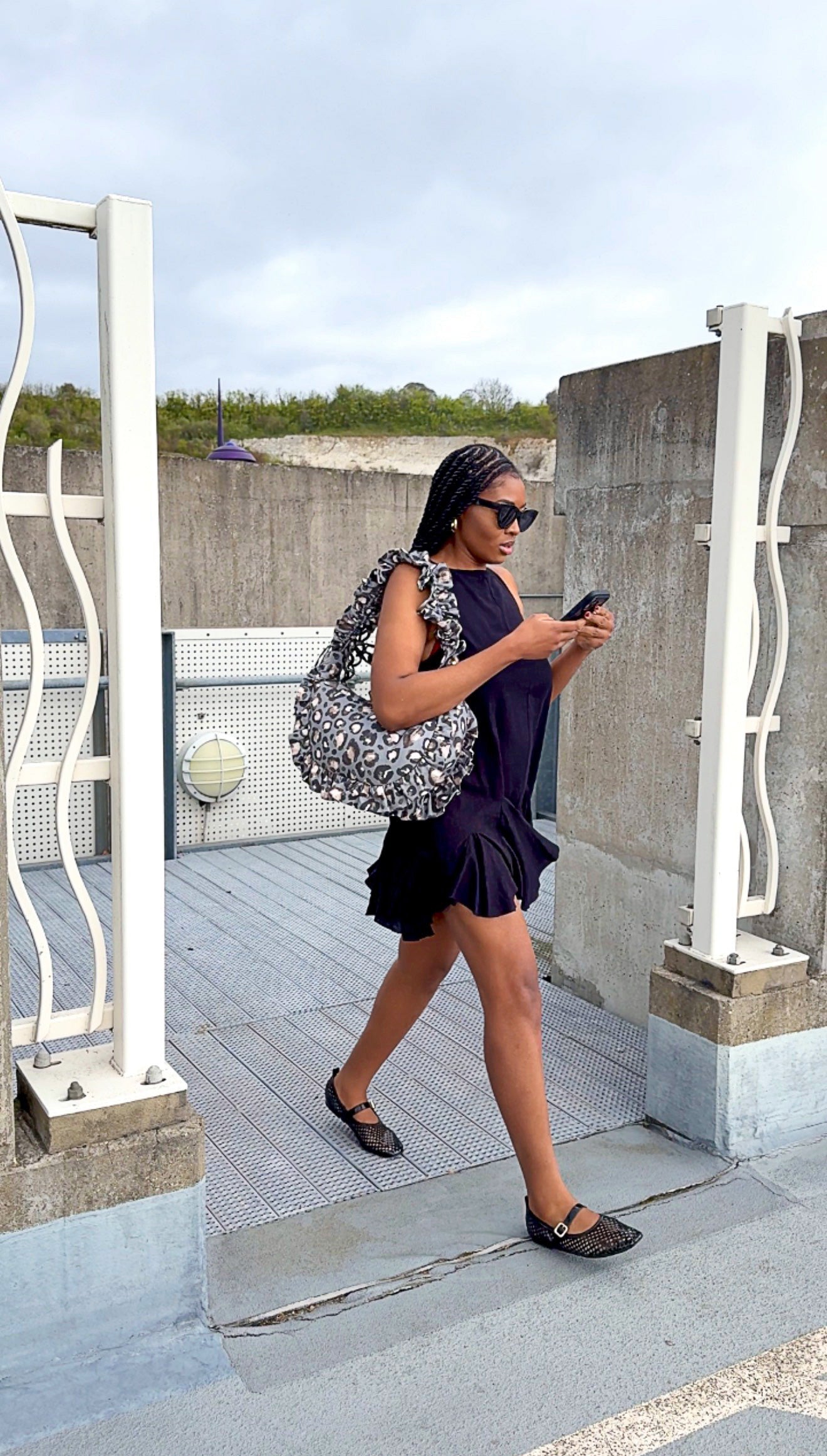 Woman in a black dress with a slate leopard print ruffled crescent bag walking on a concrete surface.