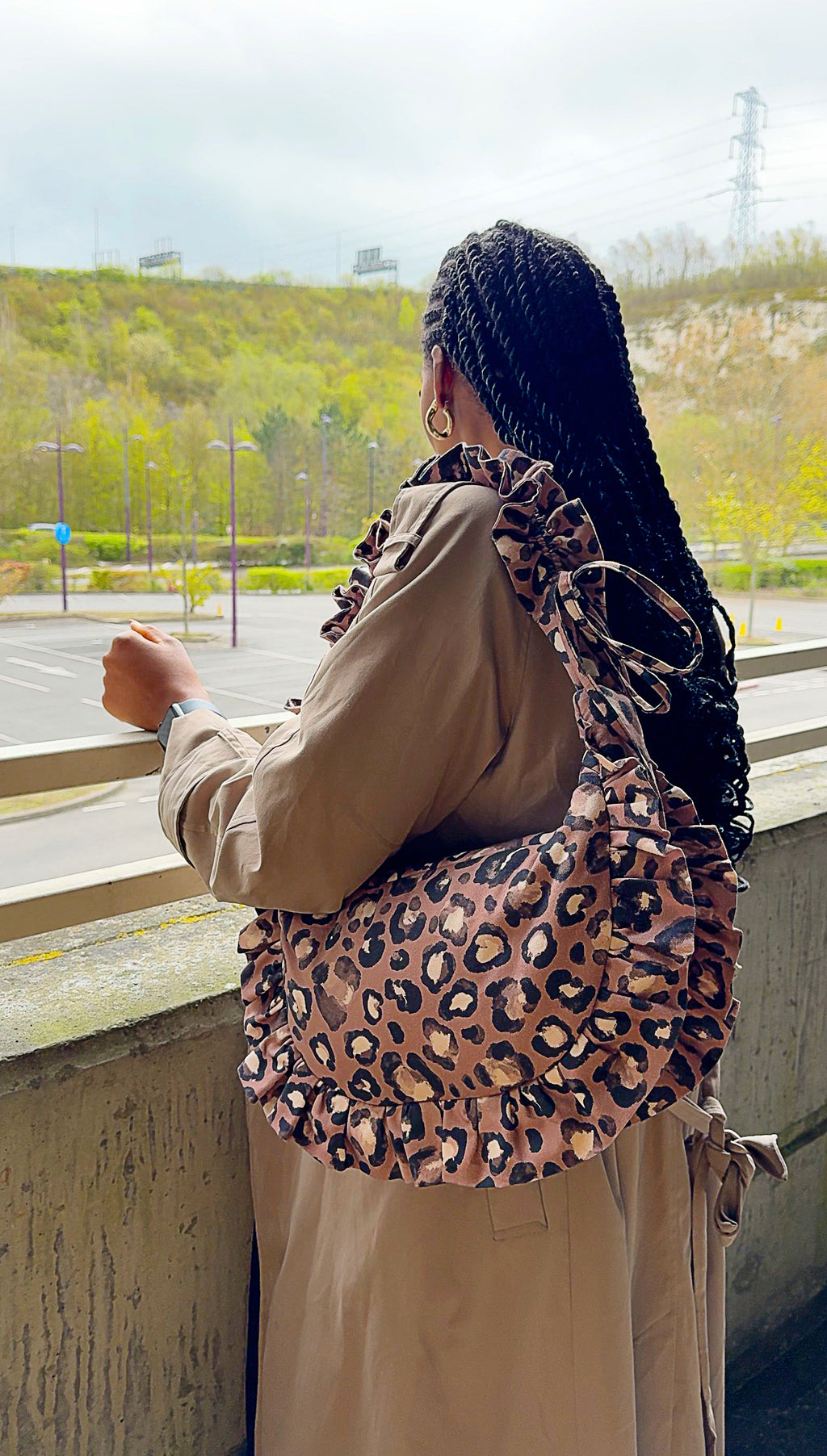 Person wearing a taupe brown ruffled crescent handbag with a scenic background
