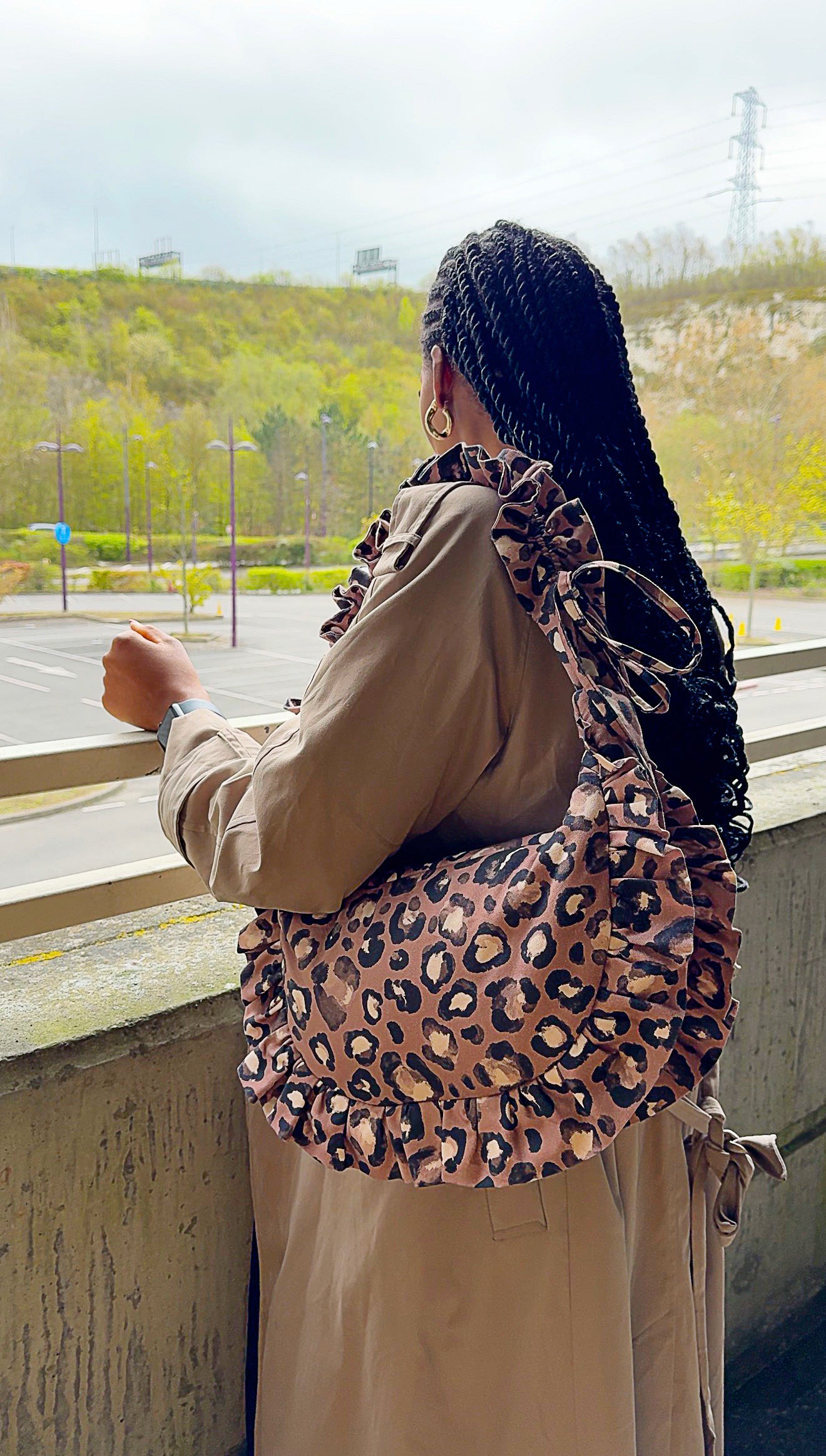 Person wearing a taupe brown ruffled crescent handbag with a scenic background