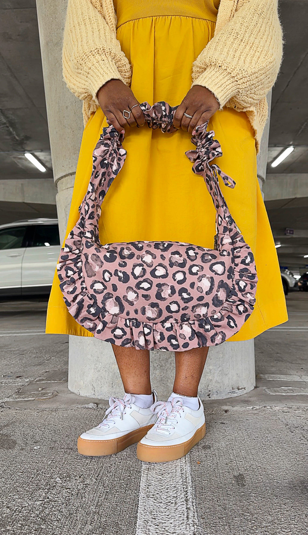 Person holding a taupe brown ruffled crescent handbag in an underground parking lot