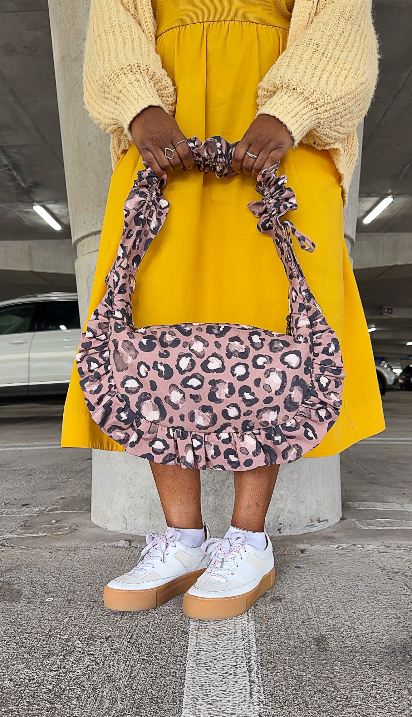 Person holding a taupe brown ruffled crescent handbag in an underground parking lot